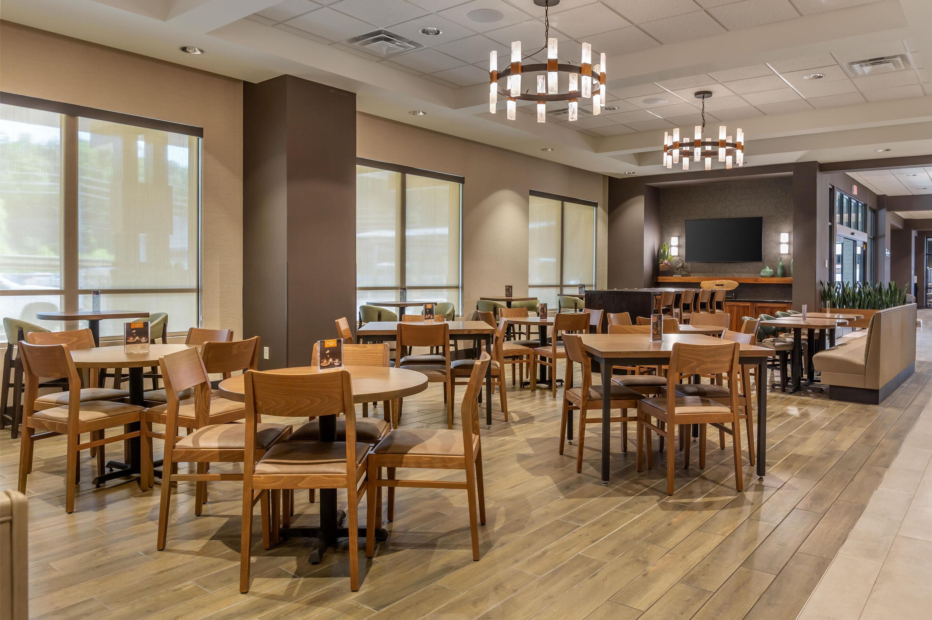 A spacious dining area with multiple wooden tables and chairs, large windows, and a modern chandelier.