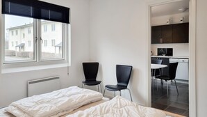 Un dormitorio con dos camas, una ventana con persianas, un radiador y vistas a una cocina y zona de comedor.
