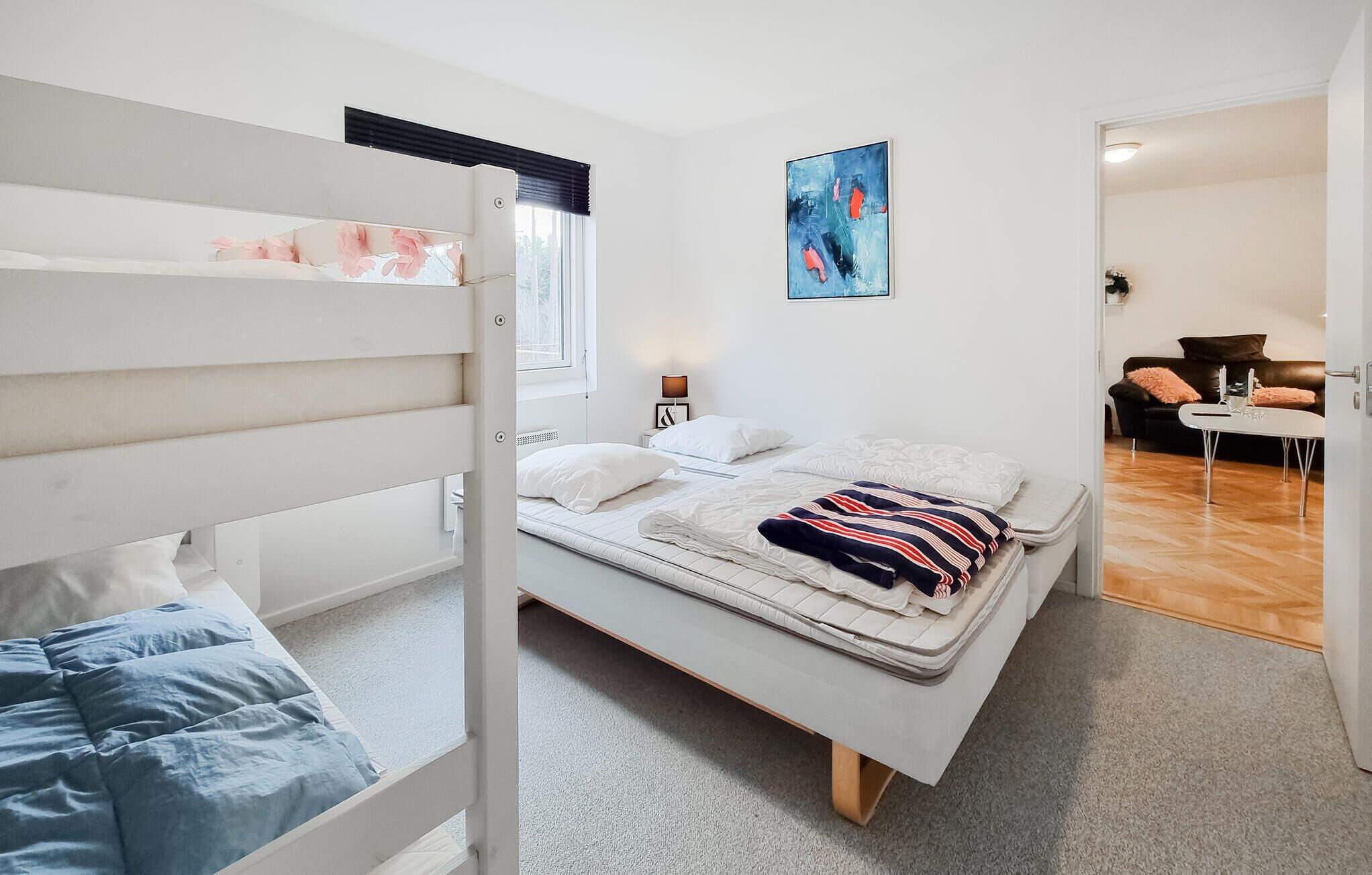 Une chambre avec un lit superposé, un lit simple, un tableau, une table de chevet, une lampe et une vue sur un espace de vie comprenant un canapé et une table.
