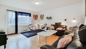 Una sala de estar moderna con un sofá de cuero negro, una mesa de centro blanca y una silla negra. Hay una ventana con persianas, un radiador y una mesita de noche con una lámpara.