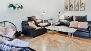 Una sala de estar moderna con dos sofás de cuero negro, una mesa de centro blanca y un sillón negro con cojines mullidos. Se observan marcos de cuadros en la pared y un pequeño estante con flores blancas.
