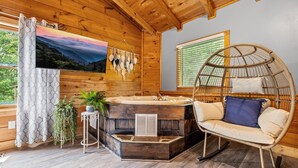 L’intérieur d’un chalet en bois, avec un plafond en bois, une grande fenêtre munie de stores, un fauteuil rond en osier, une petite table et un jacuzzi.