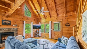 L’intérieur chaleureux d’un chalet avec une cheminée en pierre, des canapés bleus, un ventilateur de plafond et de grandes fenêtres offrant une vue sur l’extérieur.