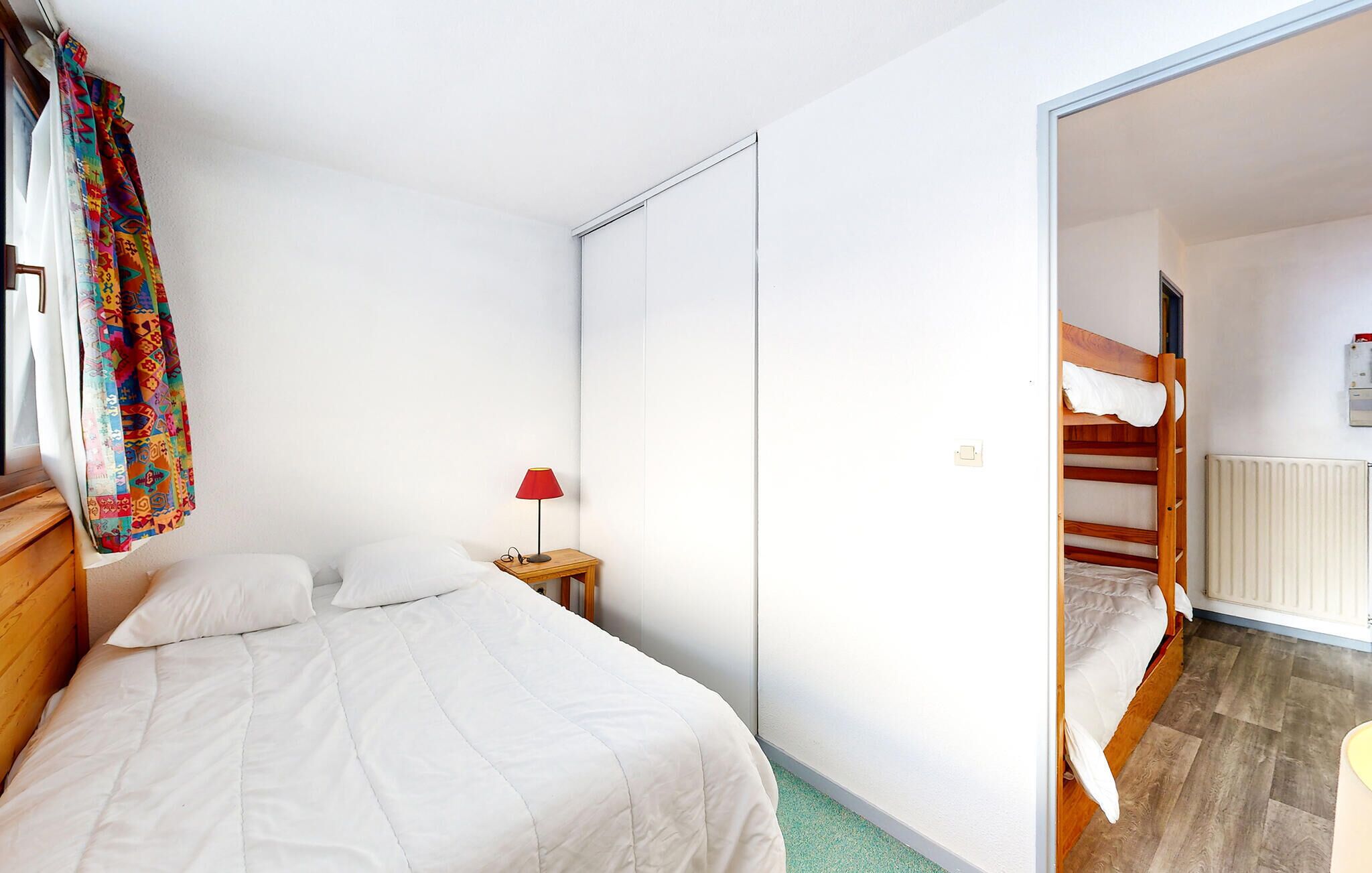 Ein Schlafzimmer mit einem Bett, einem Nachttisch mit Lampe, einem Kleiderschrank und Blick auf ein Stockbett im angrenzenden Raum.