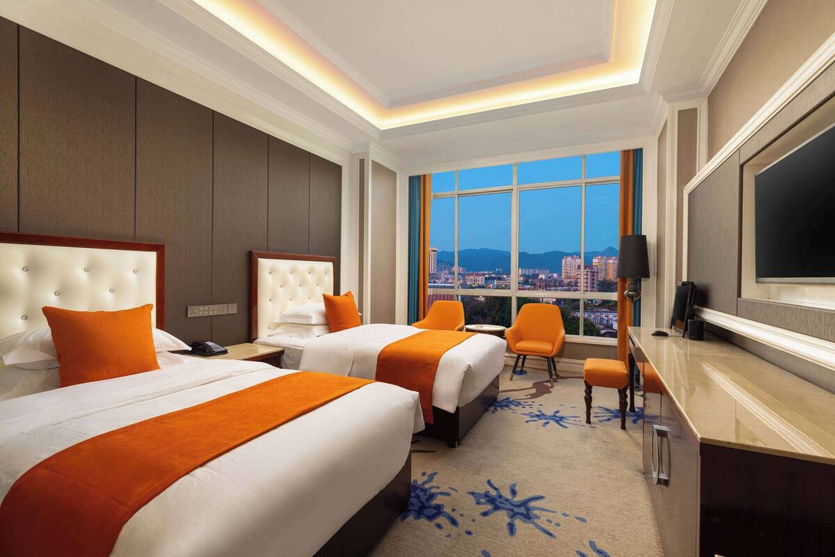 Ein Hotelzimmer mit zwei Betten, einem Schreibtisch, einem Stuhl und einem großen Fenster mit Blick auf die Stadt.