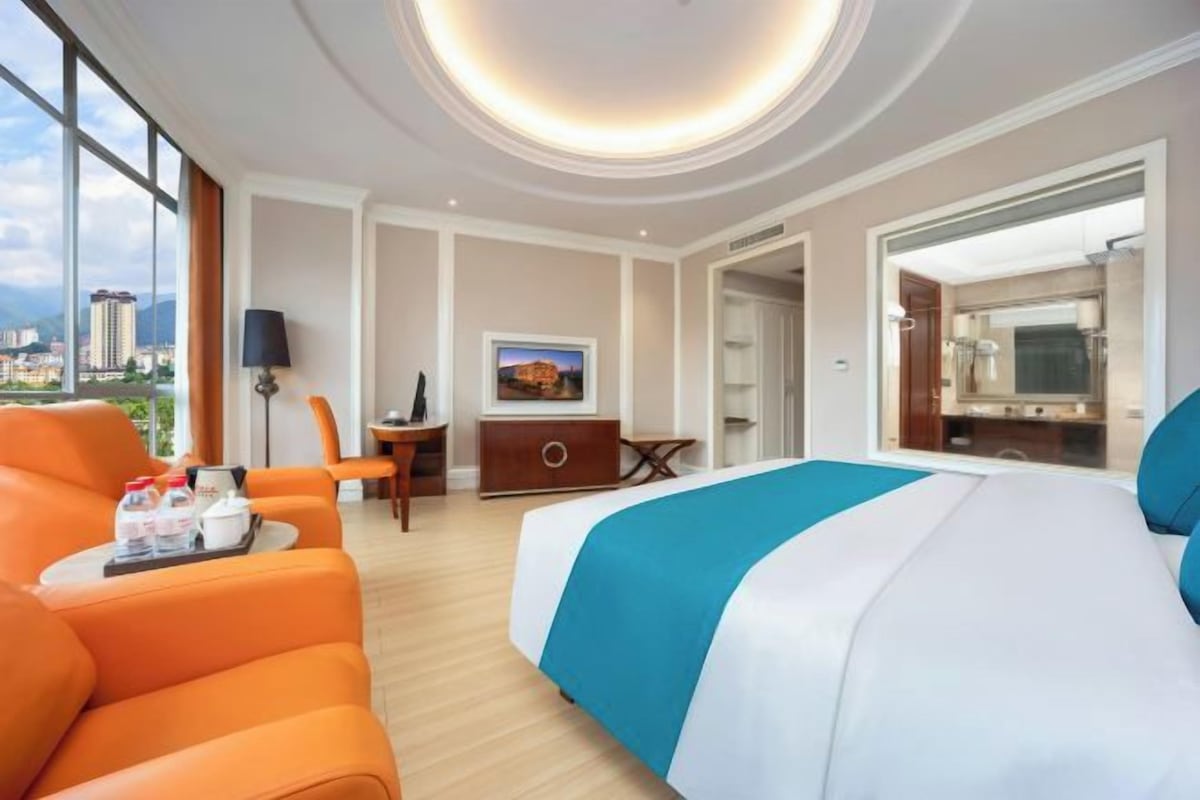 A hotel room with a large bed, a seating area with a sofa and armchairs, a desk with a chair, a television, and a view of the city.