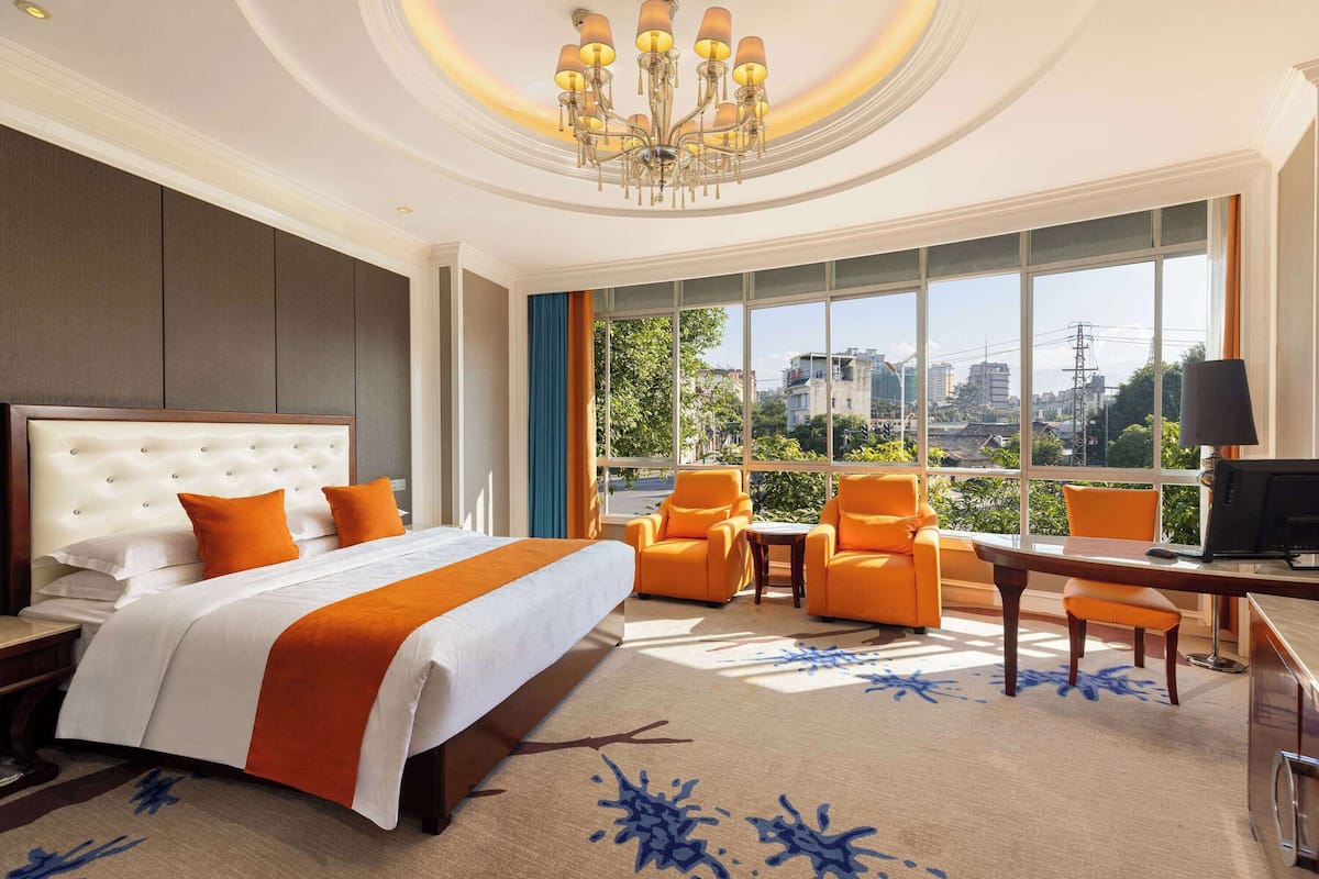 Ein Hotelzimmer mit einem großen Bett, orangenen Sesseln, einem Schreibtisch und Blick auf die Stadt durch große Fenster.
