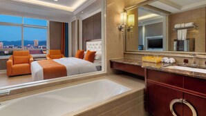 A hotel room with a large bed, two orange armchairs, a bathtub, a vanity with a mirror, and a city view.