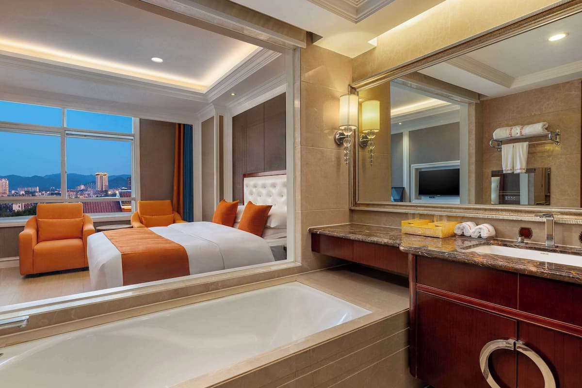 Ein Hotelzimmer mit einem großen Bett, zwei orangefarbenen Sesseln, einer Badewanne, einem Schminktisch mit Spiegel und Blick auf die Stadt.