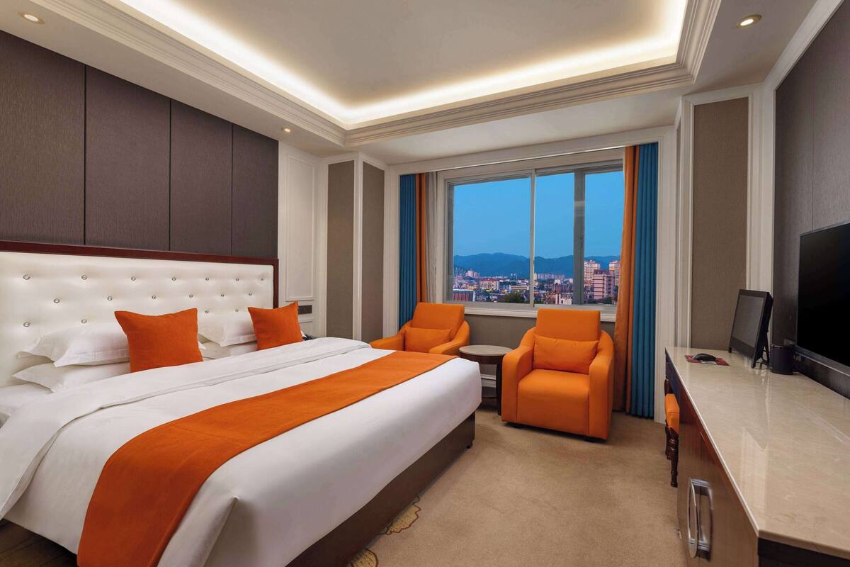 Ein Hotelzimmer mit einem großen Bett, zwei orangenen Sesseln, einem kleinen Tisch, einem Fernseher und Blick auf die Stadt durch das Fenster.
