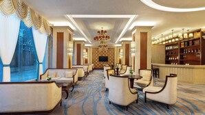 A hotel lobby with a long reception desk, multiple seating areas, a chandelier, and a large window with curtains.