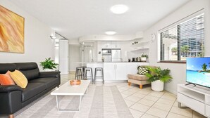 A modern living room with a black leather sofa, a coffee table, and a TV. There is a kitchen area with white cabinets and a dining space with stools. - Charming 1-Bedroom Apartment with Rooftop Pool & Prime Cairns Location (Cairns City)