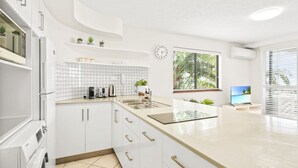 A modern kitchen with white cabinets, a sink, a countertop with appliances, and a window with a view of palm trees. - Charming 1-Bedroom Apartment with Rooftop Pool & Prime Cairns Location (Cairns City)