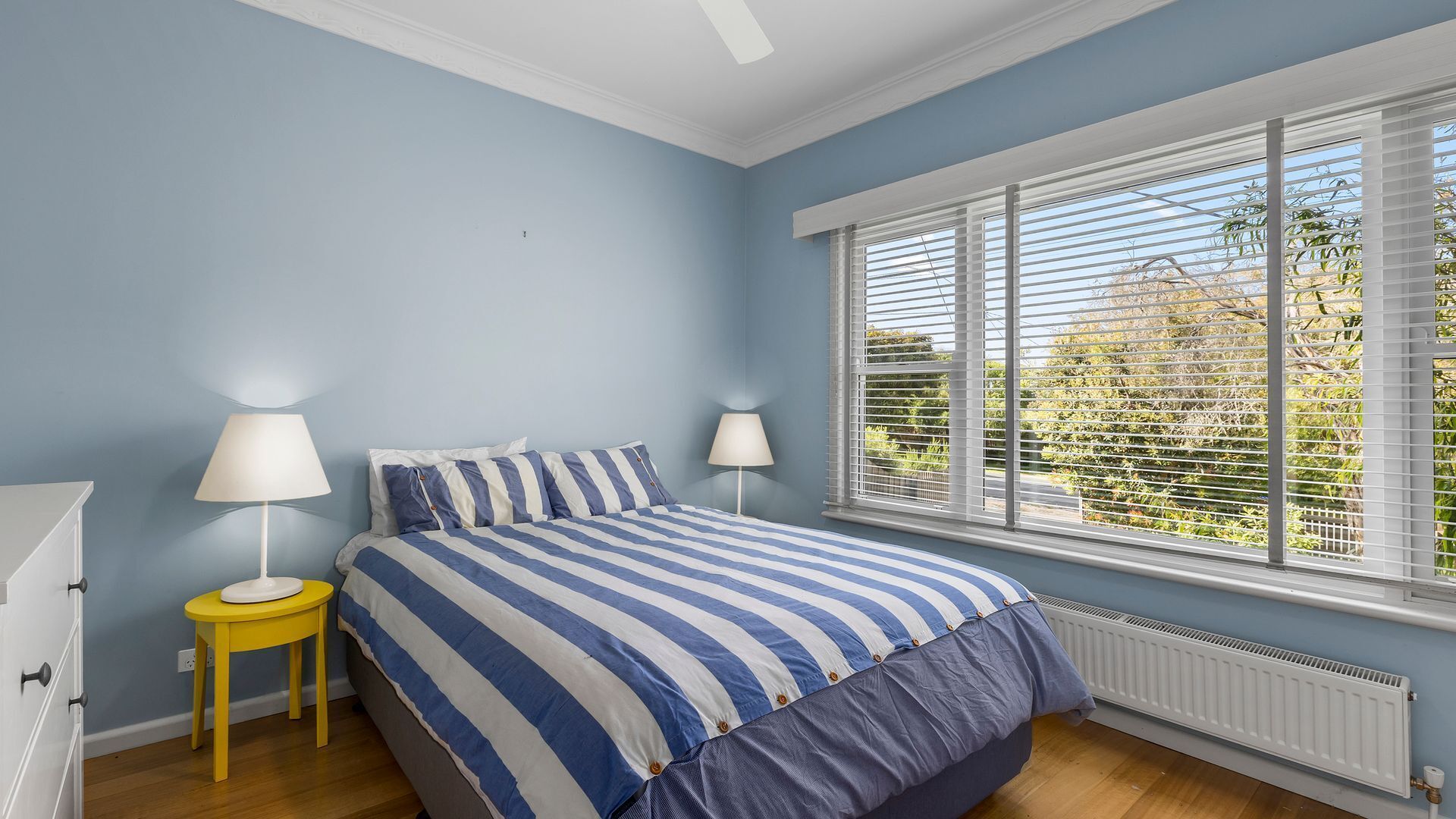 A bedroom with a bed, two bedside lamps, a window with blinds, and a radiator.