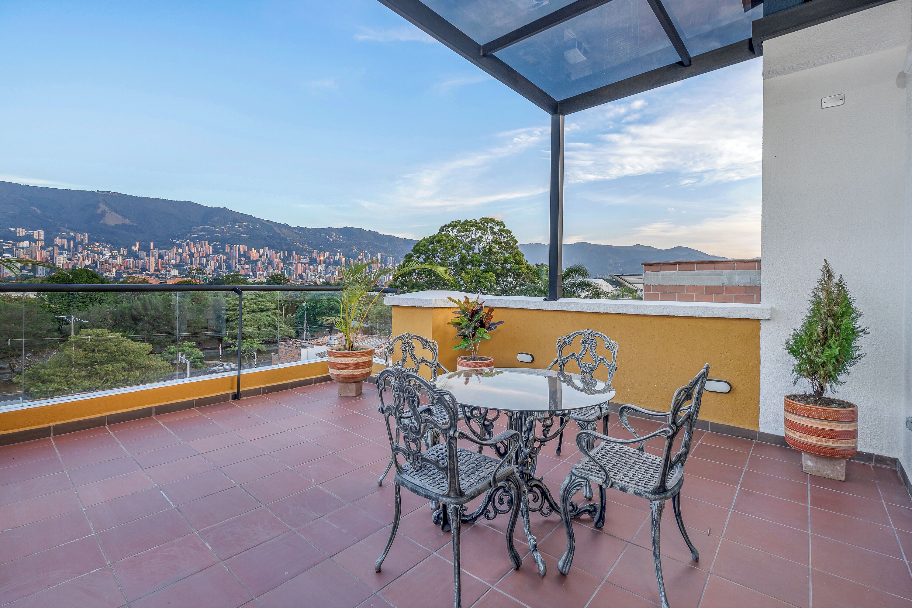 A rooftop terrace with a glass balustrade, a round table with chairs, potted plants, and a cityscape view in the background.