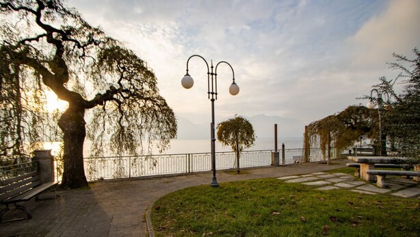 A paved walkway with benches, a lamppost, and a tree-lined path by a body of water. - [Verbania-Suna] Love Nest Apartment - 50m from the lake - Free Wi-Fi (Verbania)