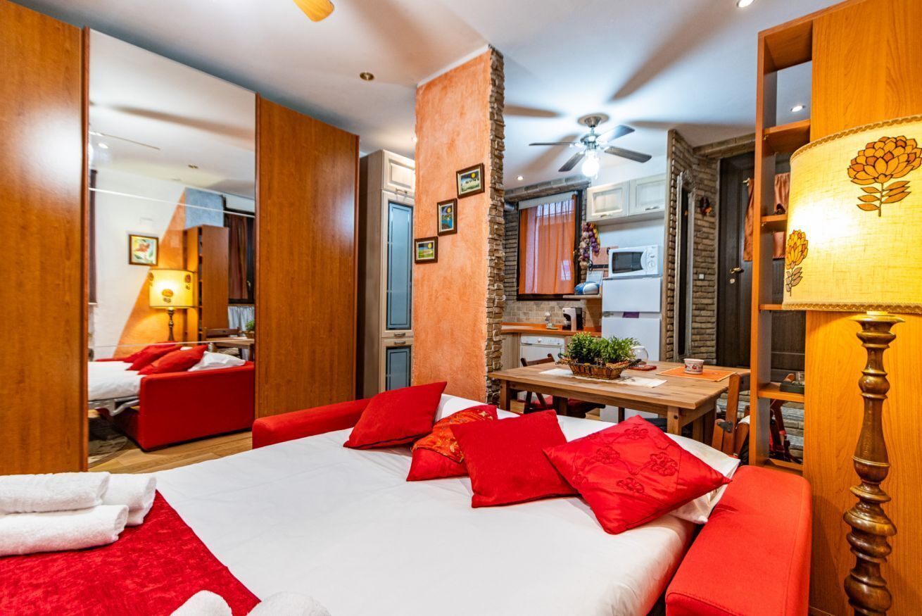 Ein Hotelzimmer mit einem Bett, roten Kissen, einem Holztisch, einem Deckenventilator und einem durchgegangene Tür erkennbaren Küchenbereich.