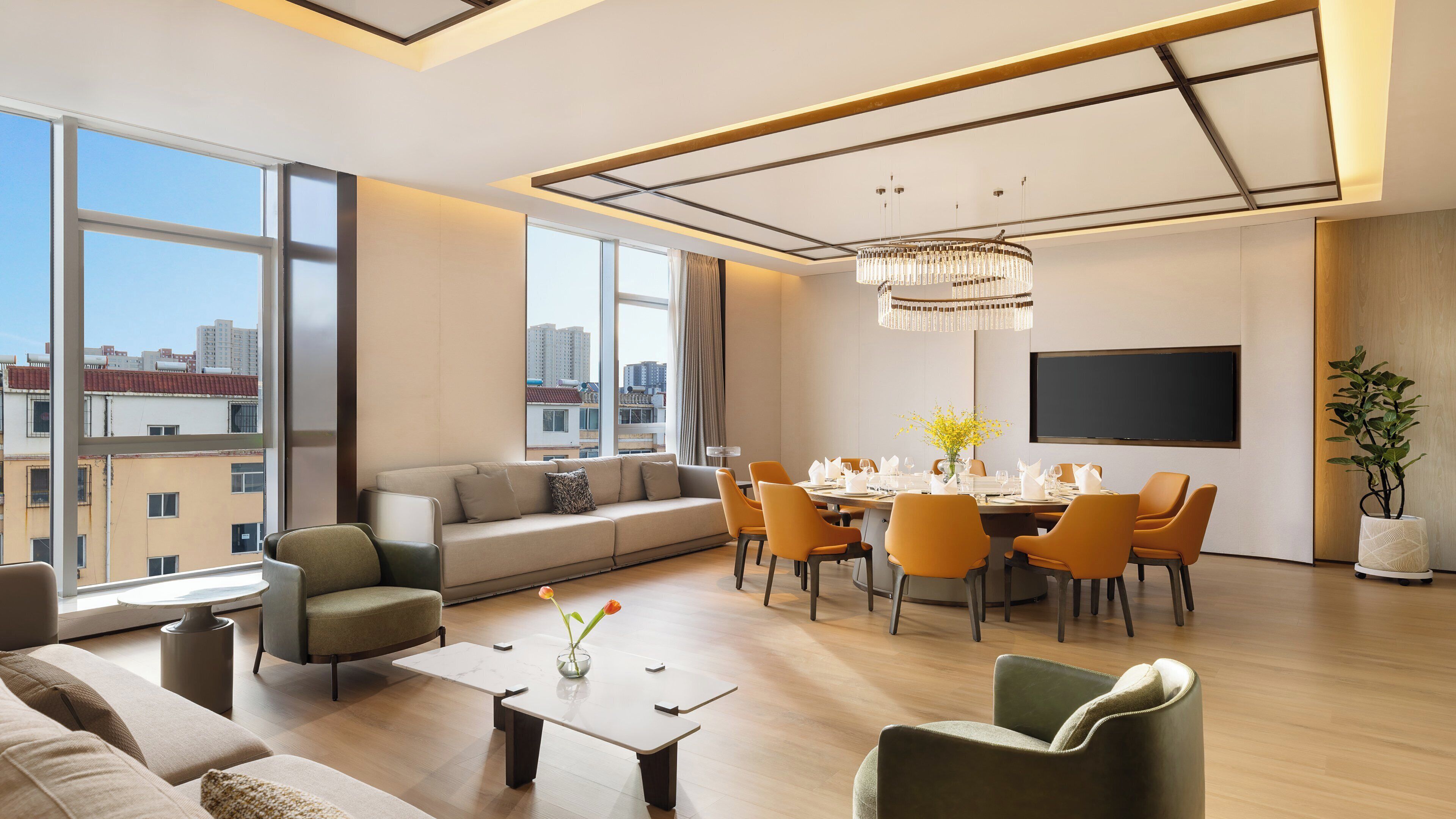 A modern living room with a dining area, large windows, a chandelier, and a flat-screen TV.