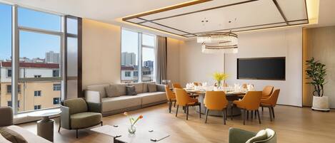 A modern living room with a dining area, large windows, a chandelier, and a flat-screen TV.