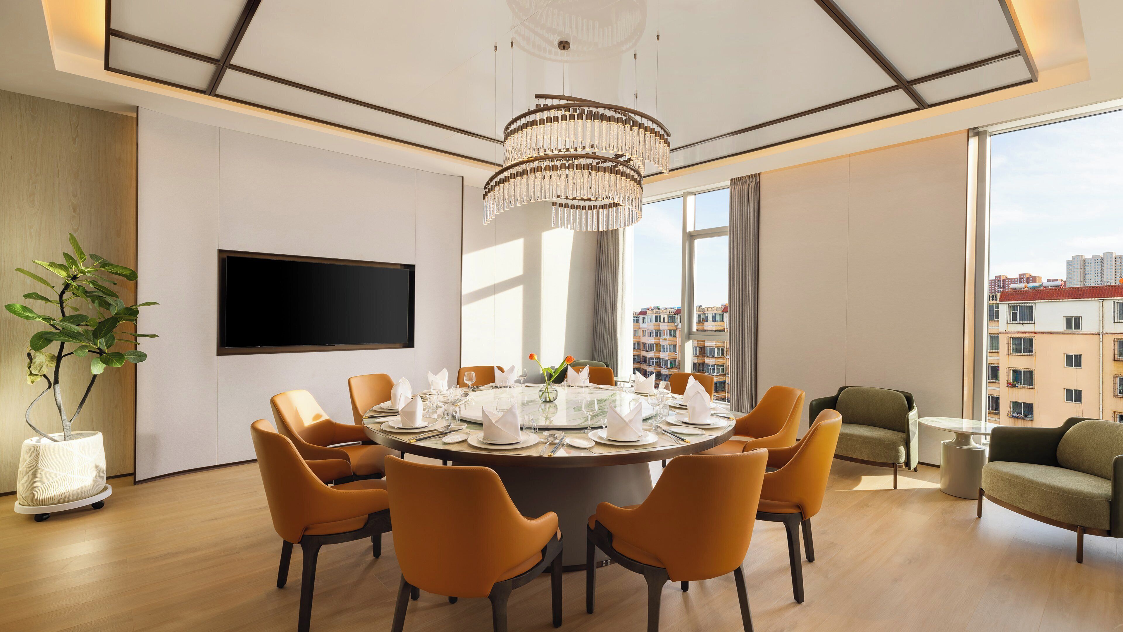 Un comedor moderno con una mesa redonda preparada para seis personas, rodeada de sillas naranjas, una lámpara de araña grande y un televisor de pantalla plana en la pared.