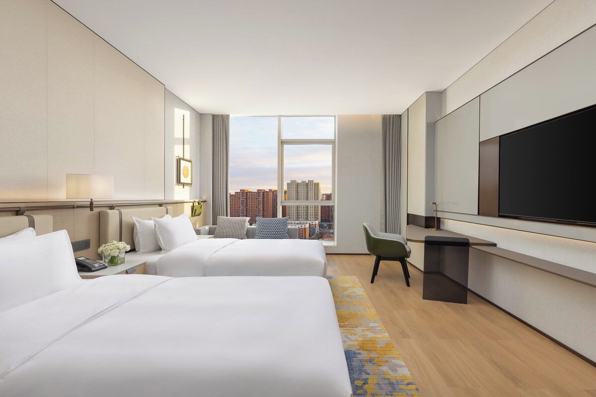 A modern hotel room with two beds, a desk, a flat-screen TV, and a city view.