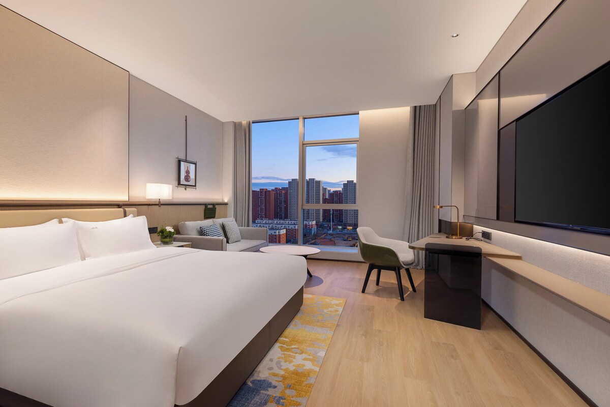 A modern hotel room with a large bed, a desk, a chair, and a large window offering a city view.