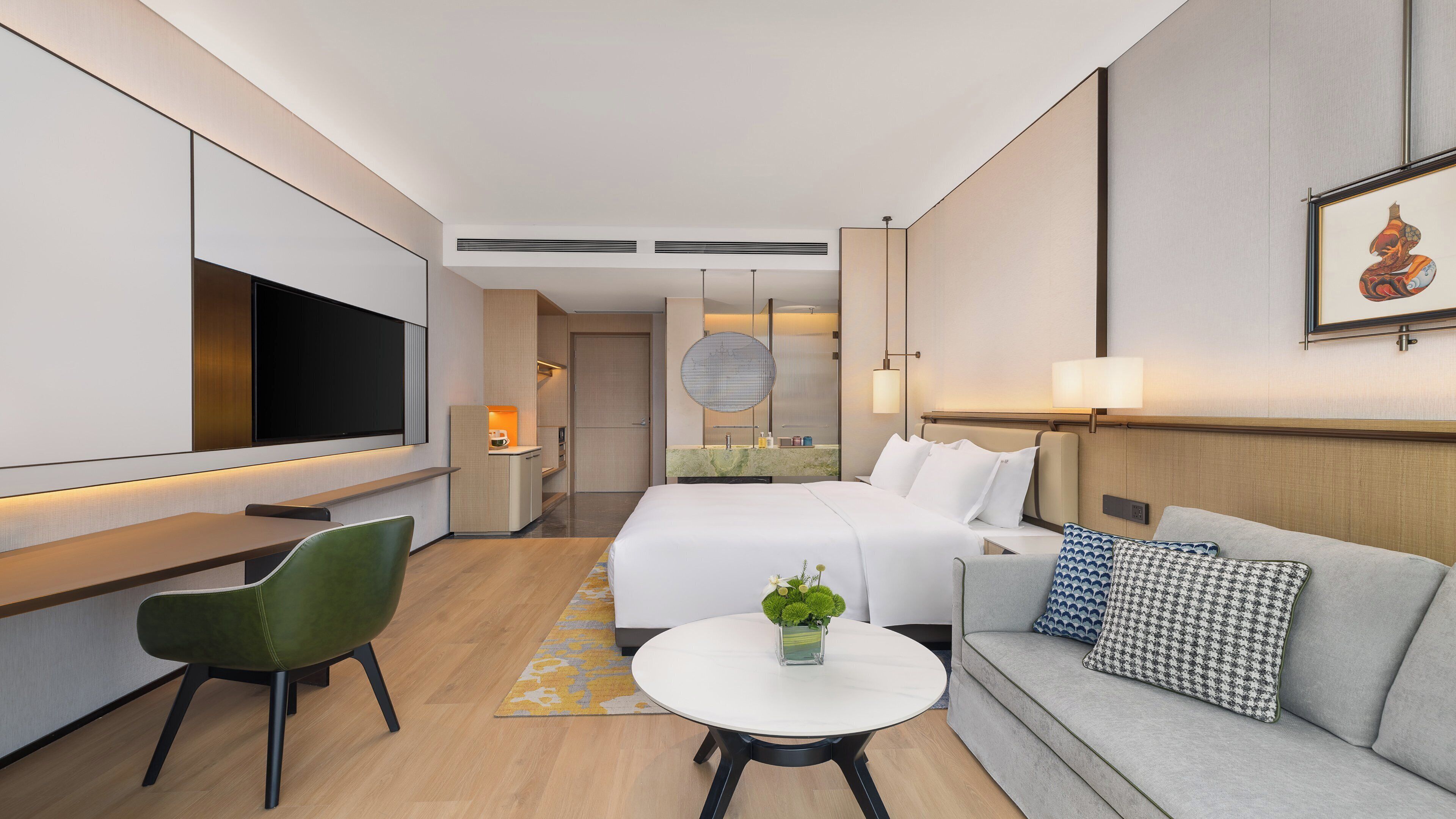 A modern hotel room with a bed, a sofa, a dining table, a TV, and a bathroom visible through an open door.