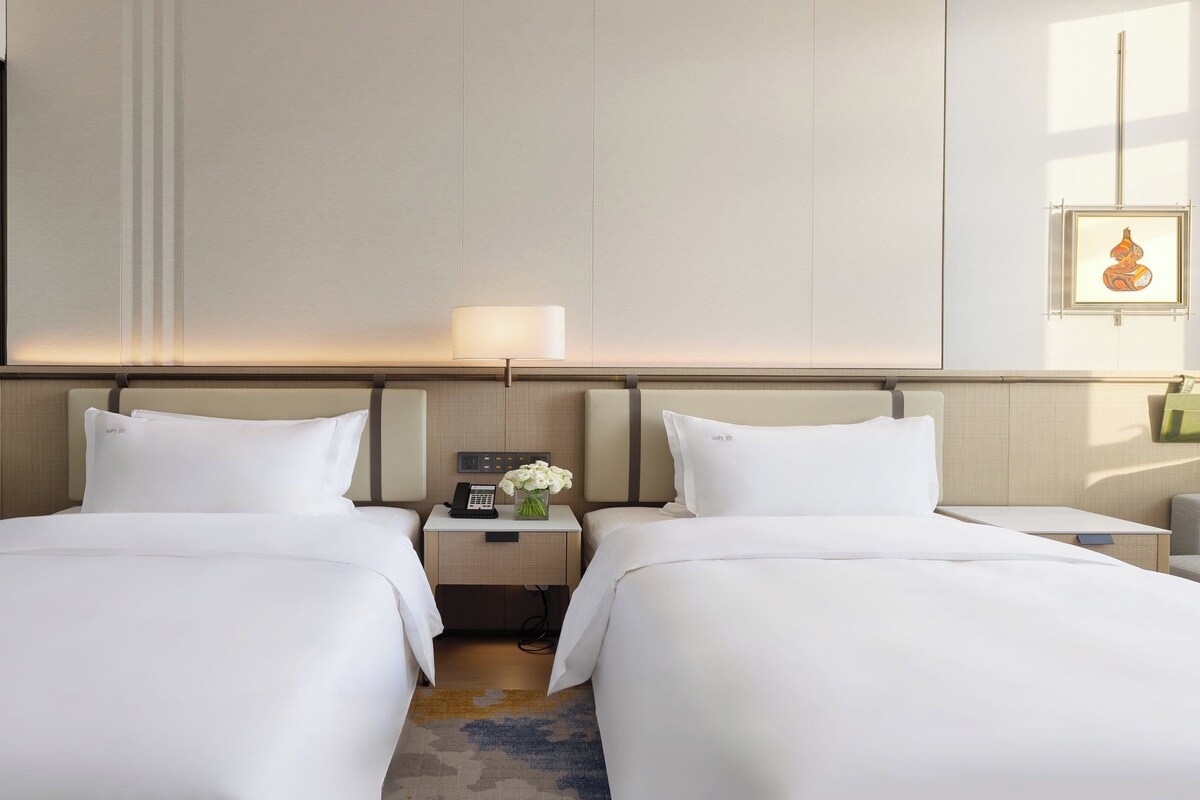 A hotel room with two beds, a nightstand with a lamp, a phone, and a vase of flowers.