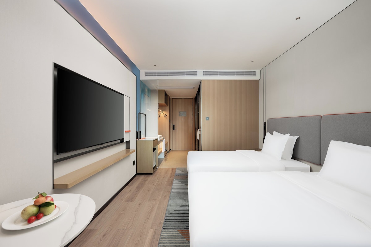 A modern hotel room with a large flat-screen TV, a bed, a bedside table, and a small seating area with a chair and a table.