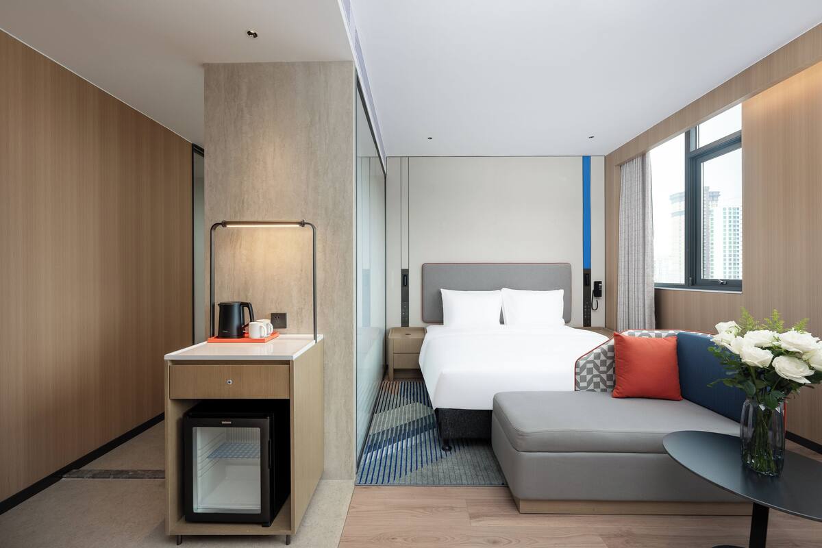 A modern hotel room with a bed, a desk with a coffee maker, a mini-fridge, a sofa, and a vase with flowers.