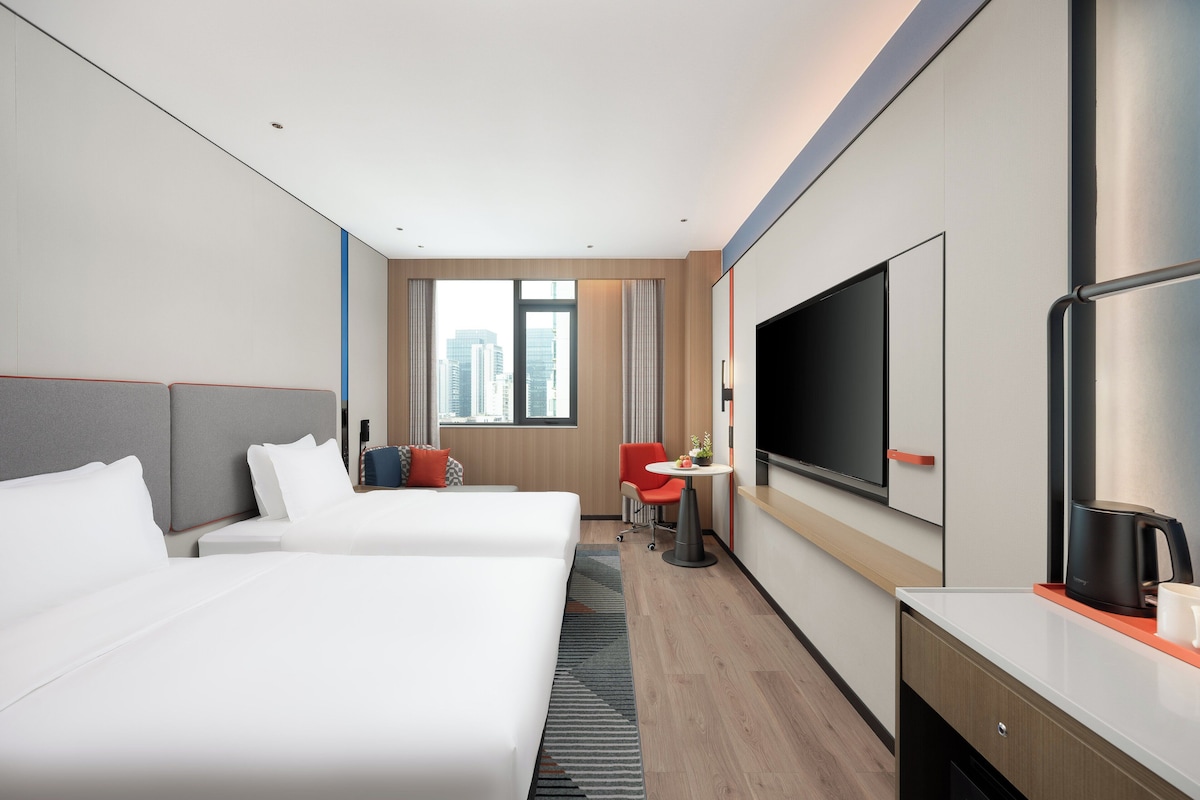 A hotel room with two beds, a desk, a TV, and a city view.