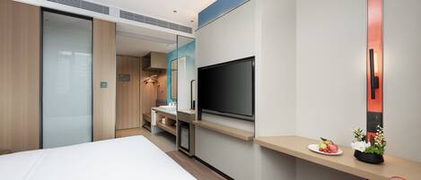 A modern hotel room with a large bed, a desk with a flat-screen TV, a small kitchenette, and a sliding door closet.