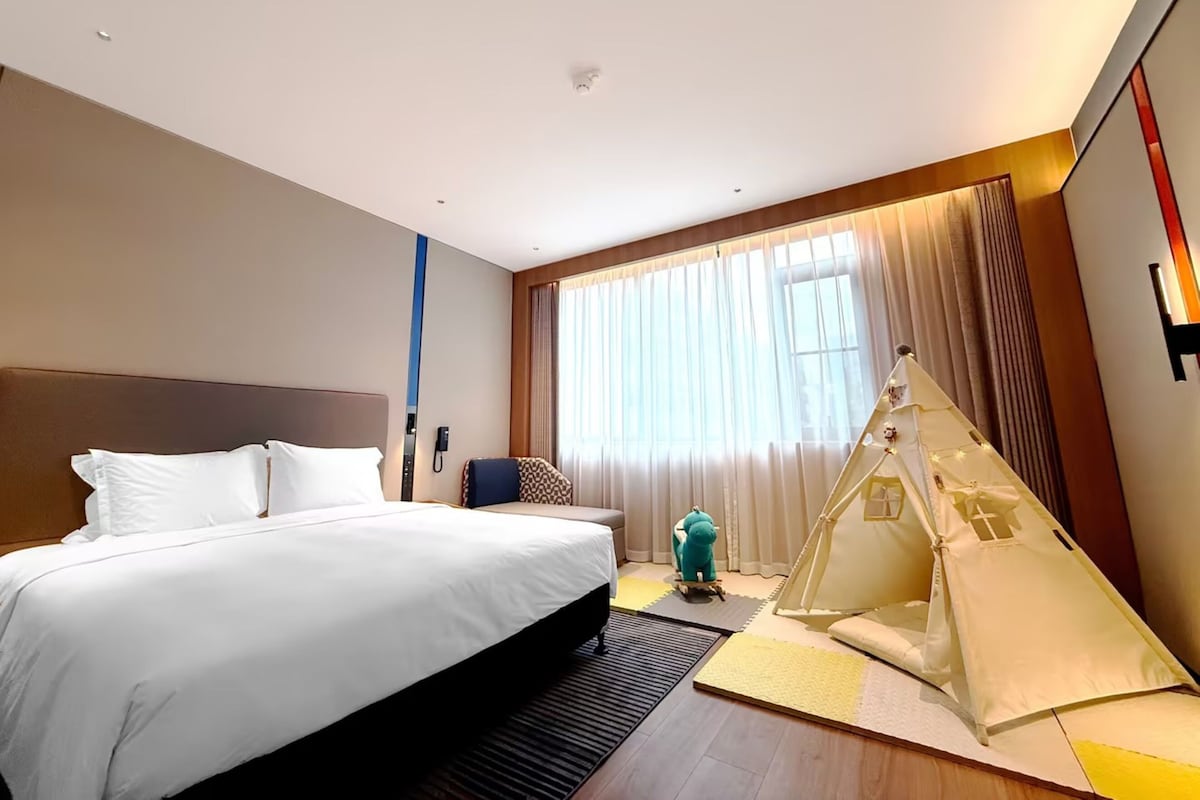A hotel room with a large bed, a small desk, a chair, a tent-like structure, and a large window with curtains.