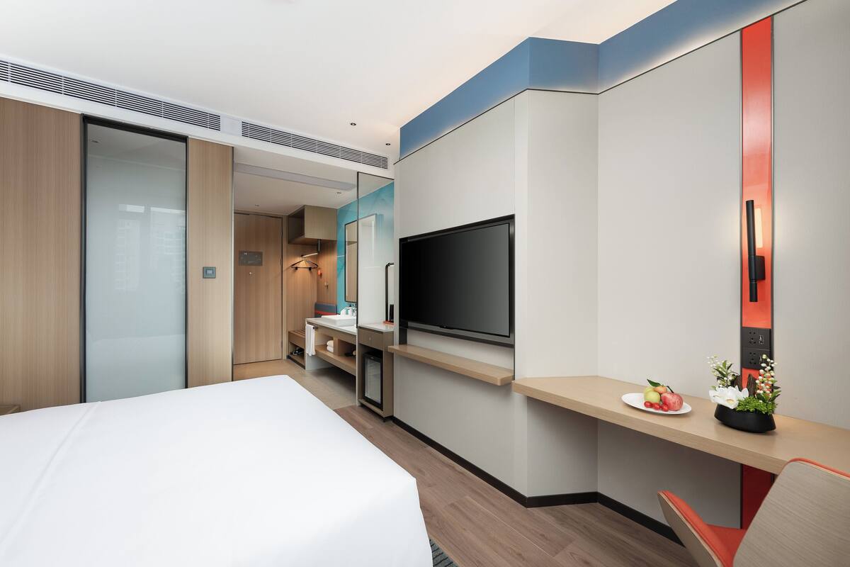A modern hotel room with a large bed, a desk with a flat-screen TV, a small kitchenette, and a sliding door closet.
