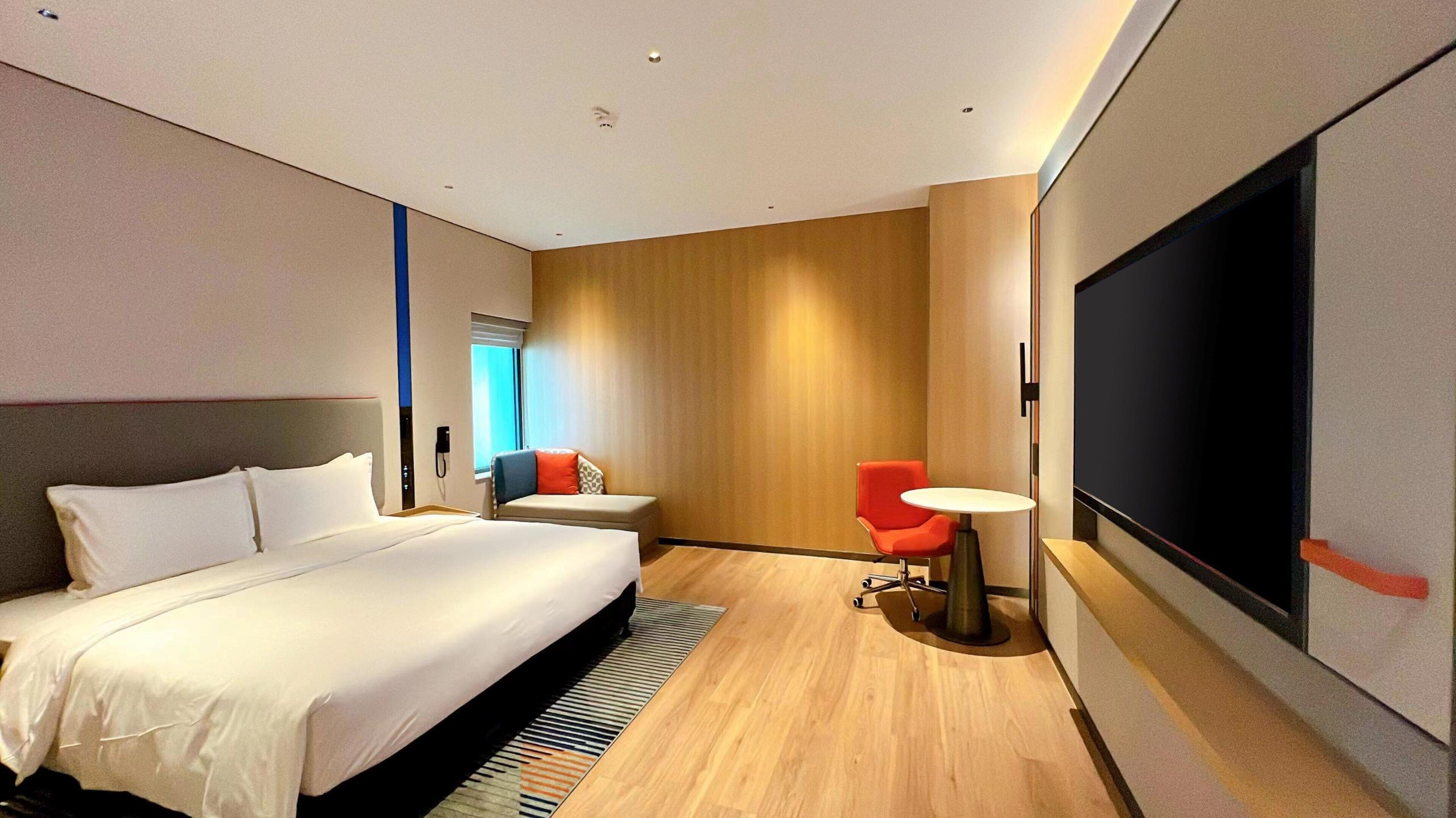 Habitación de hotel moderna con una cama grande, un sofá pequeño, una silla roja, una mesa redonda y una pantalla de televisión grande.