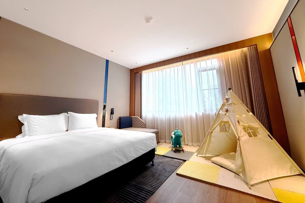 A hotel room with a large bed, a small desk, a chair, a tent-like structure, and a large window with curtains.