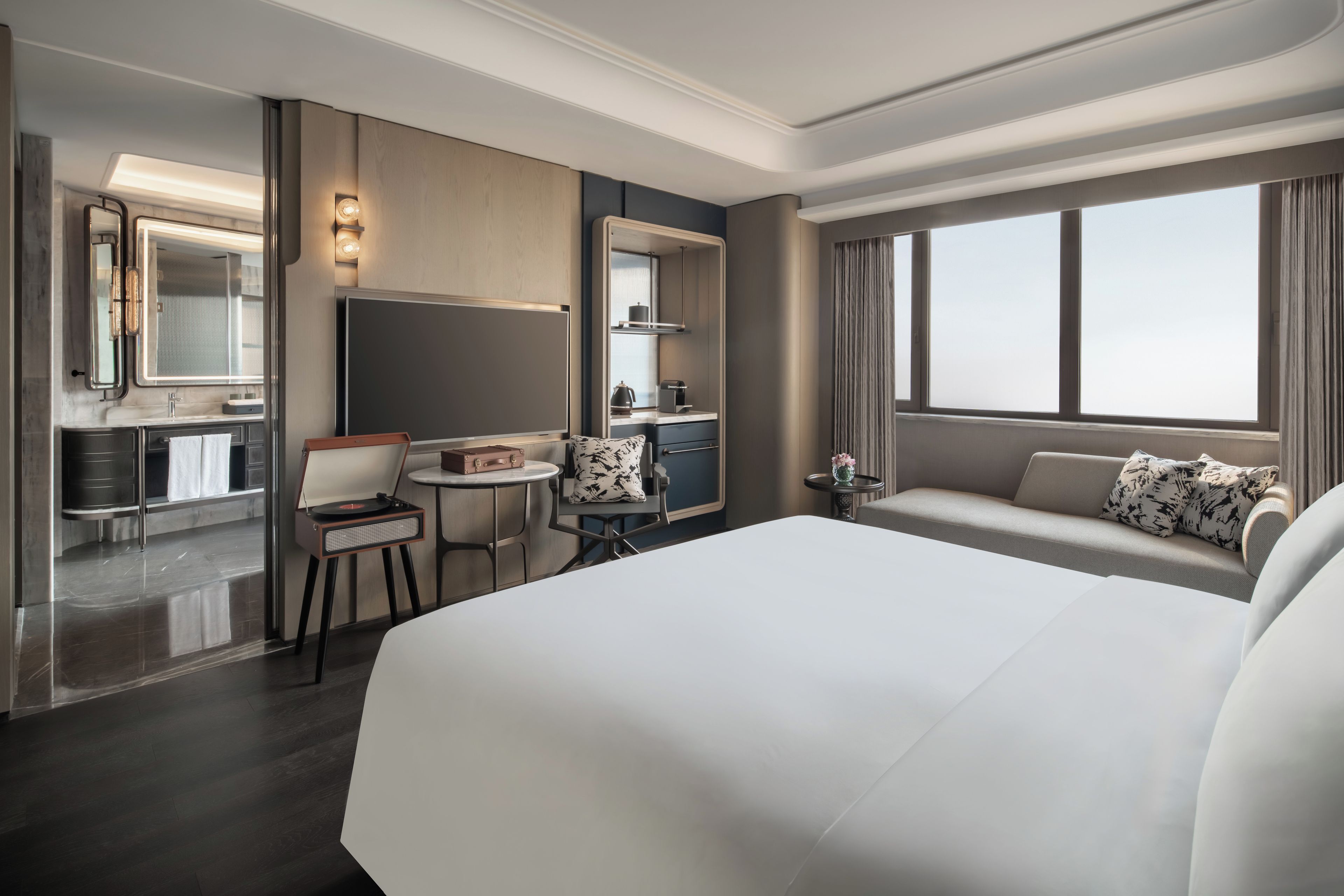 A hotel room with a large bed, a desk with a chair, a small table, a sofa, a large window, and a mirror.