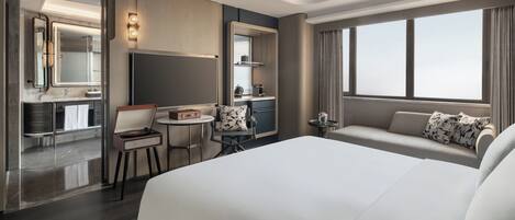 A modern hotel room with a large bed, a sitting area with a sofa and armchairs, a small round table with a chair, a vanity with a mirror, and a kitchenette.