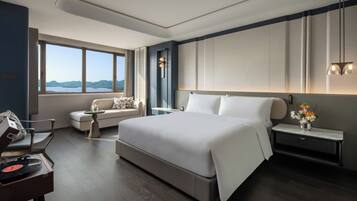 A modern hotel room with a large bed, a seating area with a sofa and armchair, a desk with a chair, and a view of the mountains through the window.