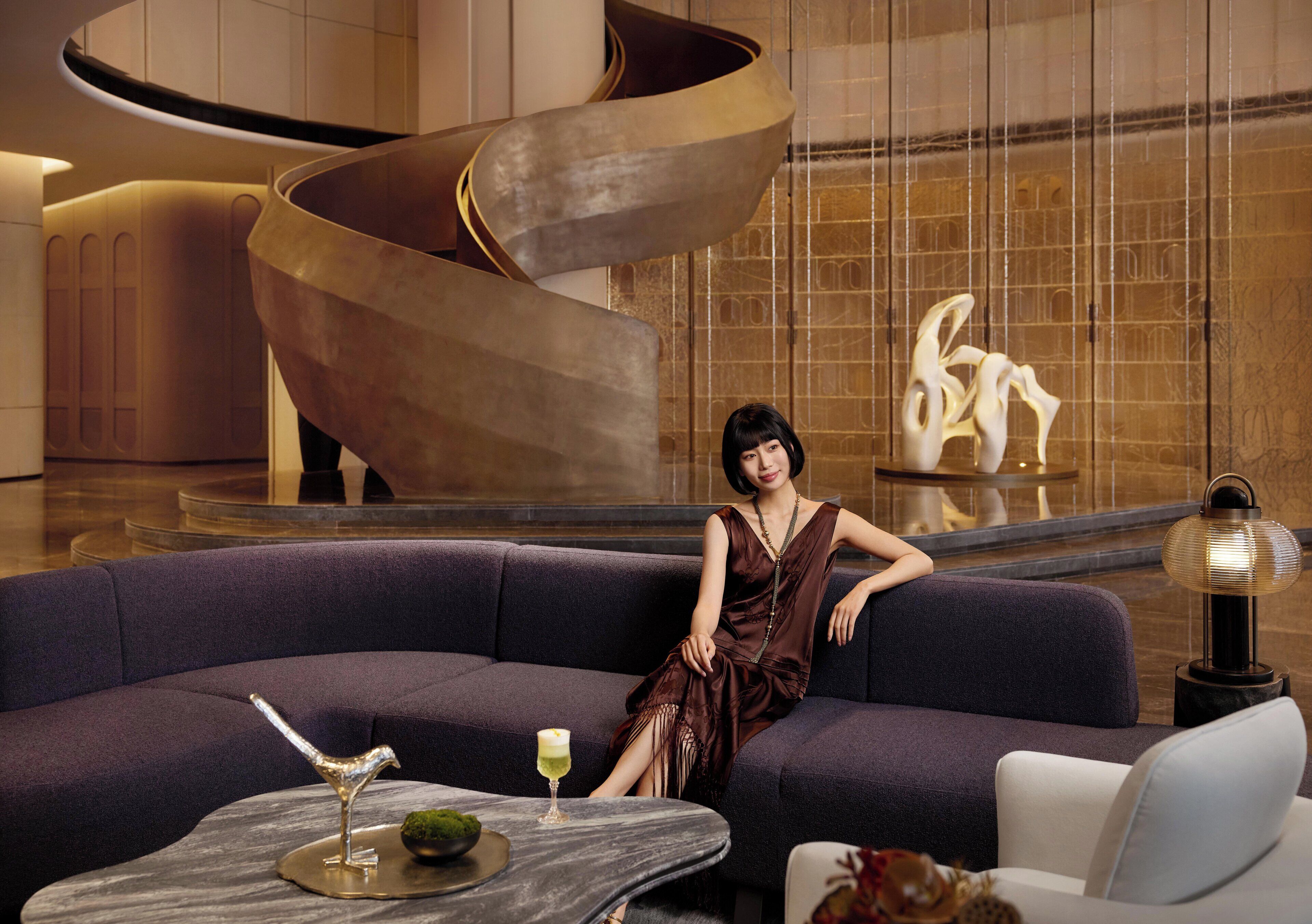 A woman sitting on a dark blue sofa in a modern lobby with a spiral staircase and a decorative sculpture in the background.