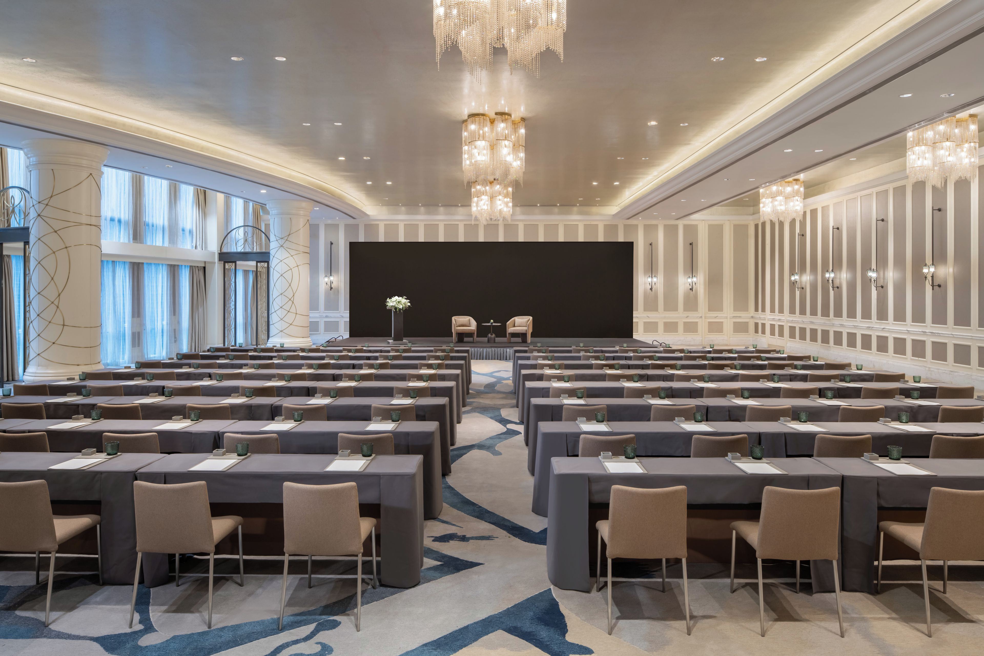 Un salón de conferencias con filas de mesas y sillas, un escenario y candelabros.