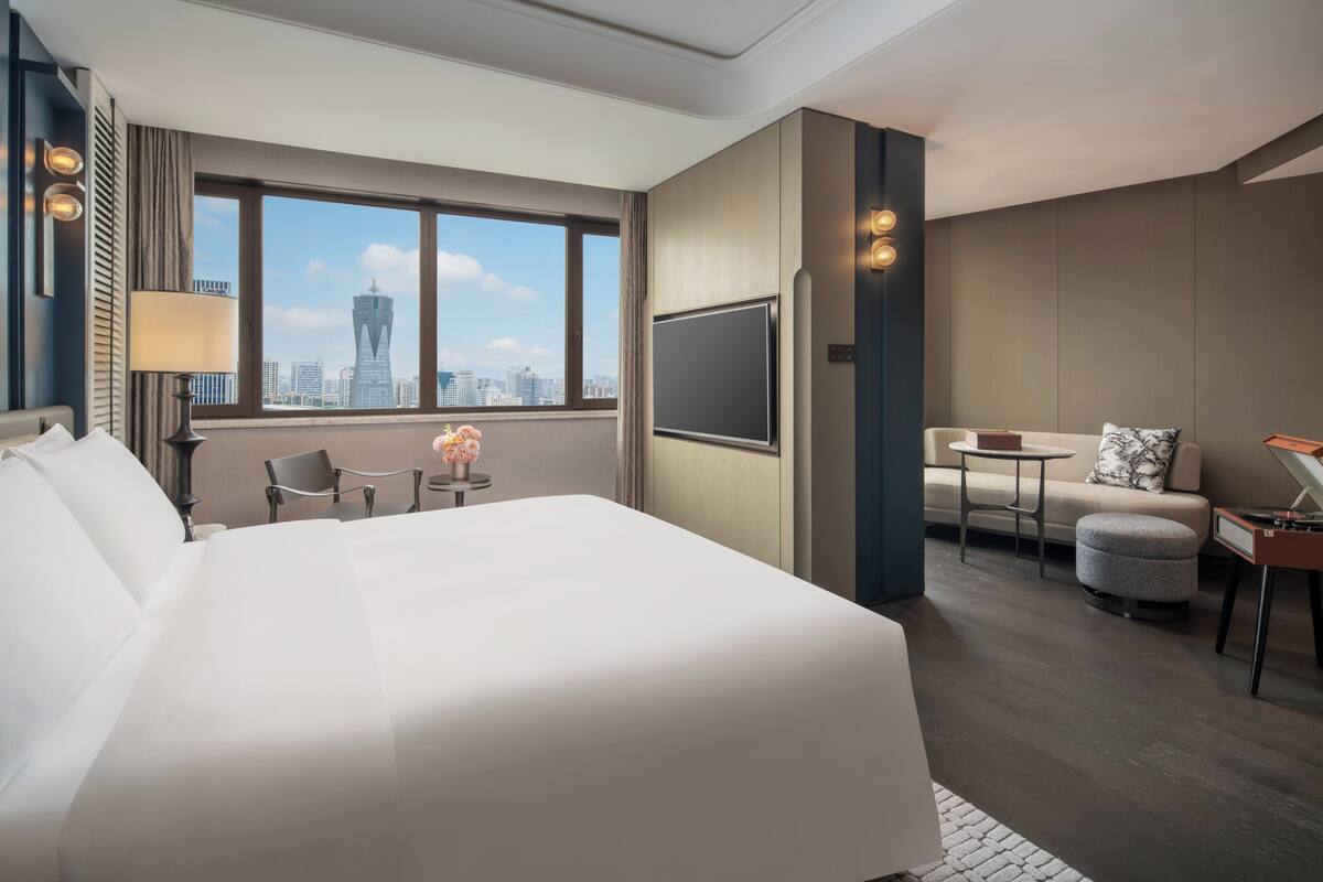 A modern hotel room with a large bed, a desk, a chair, and a sofa. There is a large window with a city view.