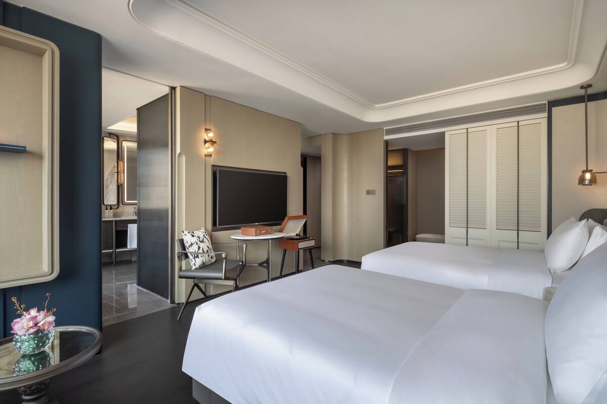 A hotel room with two beds, a desk, a chair, a TV, and a bathroom visible through an open door.
