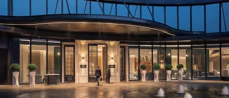 Una entrada de hotel moderna con fachada de vidrio curvada, un elemento acuático en primer plano y un interior bien iluminado.