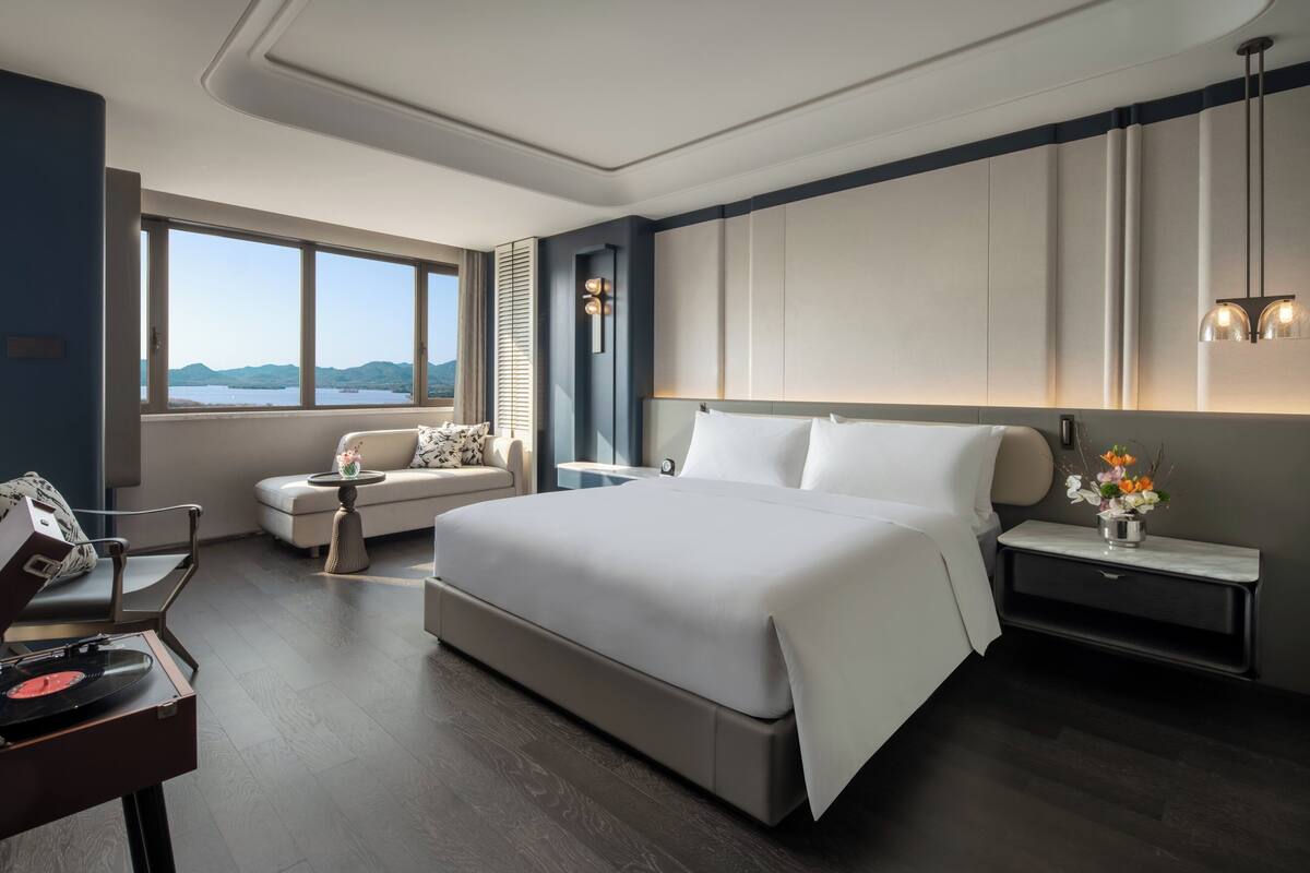 A modern hotel room with a large bed, a sofa, a desk with a record player, and a view of the mountains through the window.
