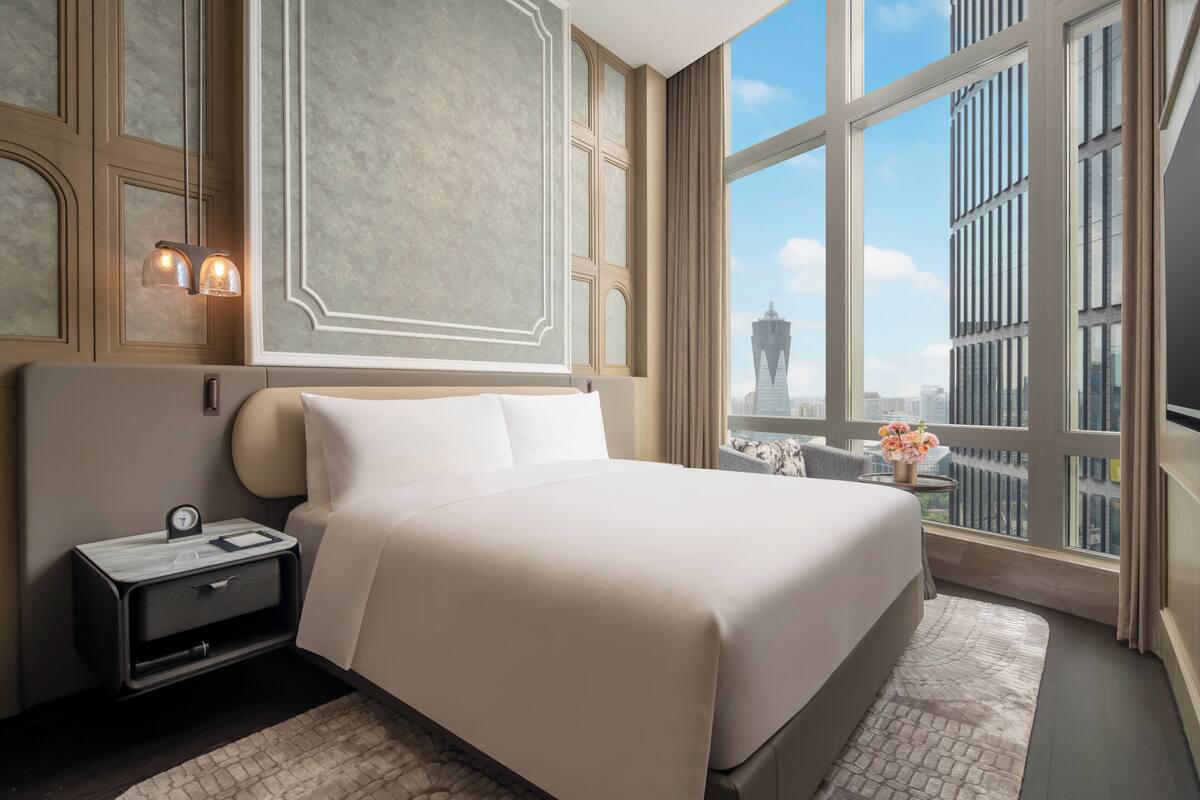 A modern hotel room with a large bed, a nightstand, a view of the cityscape, and a decorative wall panel.