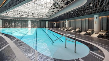 A large indoor swimming pool with a modern design, surrounded by lounge chairs and a high ceiling with structural beams.