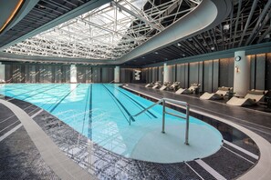 Une grande piscine intérieure au design moderne, entourée de chaises longues et dotée d’un haut plafond avec des poutres apparentes.
