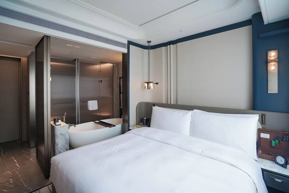 A hotel room with a large bed, a bathroom with a bathtub, and a nightstand with a clock.