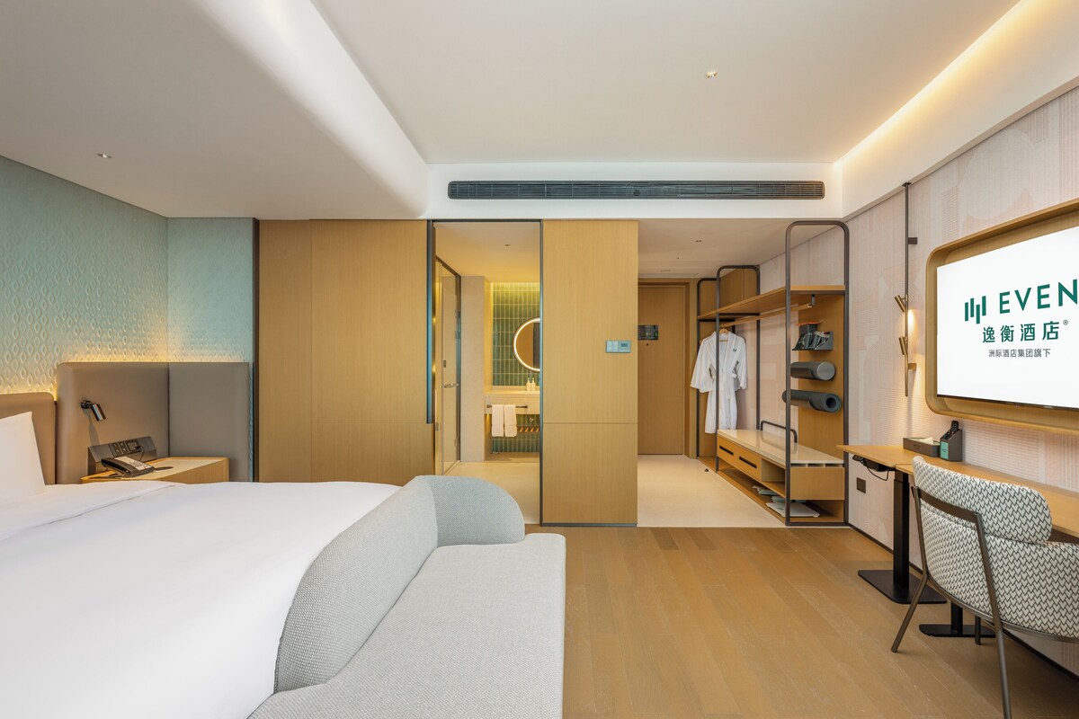 A hotel room with a bed, a desk, a chair, a wardrobe, and a bathroom visible through an open door.